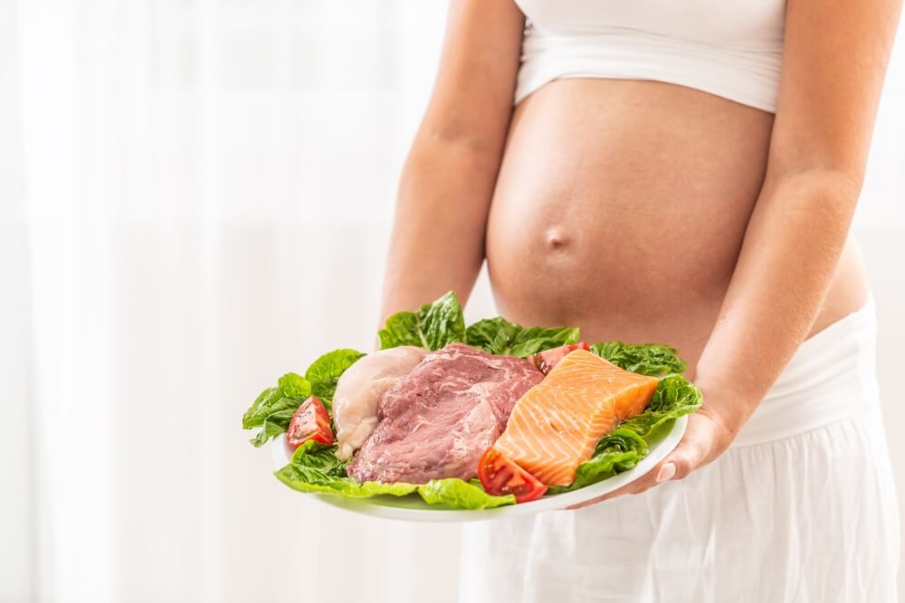 אישה בהריון מחזיקה צלחת עם בשר, עוף ודג סלמון על סלט חסה