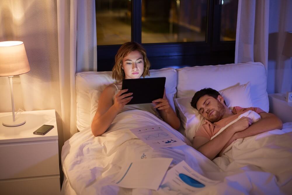 אישה קוראת מהטאבלט במיטה. בן זוגה ישן לצידה