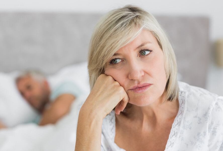 אישה בגיל המעבר יושבת על המיטה וברקע בעלה ישן