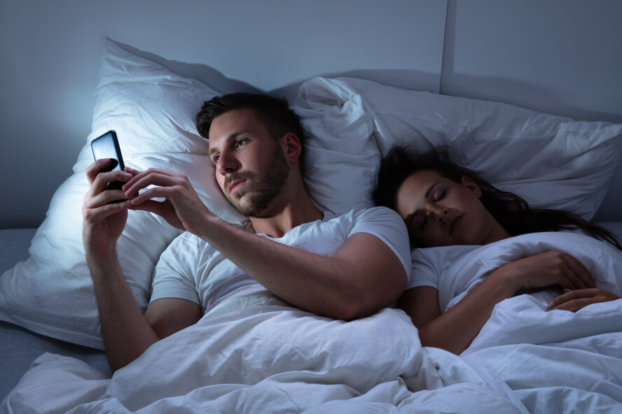 בגידה רגשית (מיקרו בגידה) - גבר משתמש בסמארטפון במיטה, לצד בת זוגו הישנה